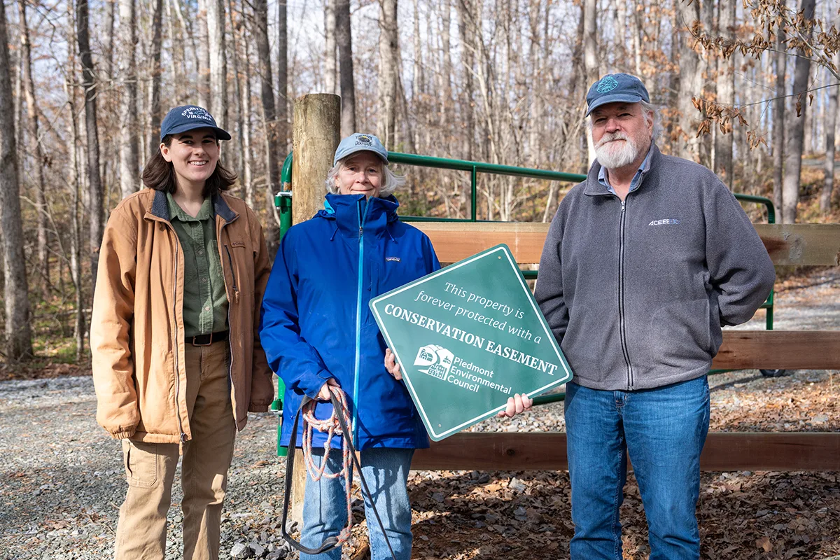 a couple holds an easement sign, standing with a PEC field rep, on a forested property in autumn