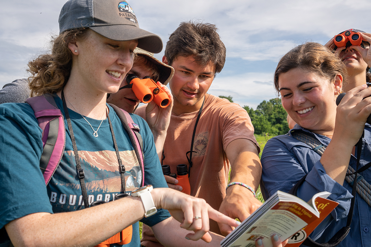 a group of young adults look at a bird identification book in a field