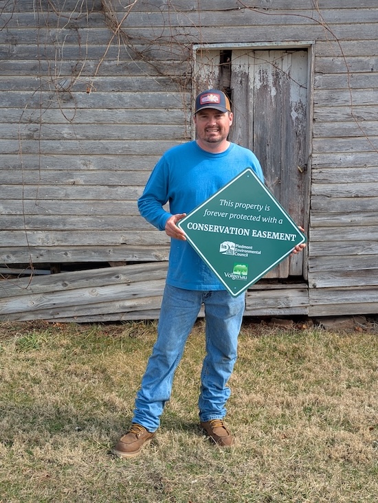 A man standing in front of a barn holding a green sign that reads "This property is forever protected with a conservation easement"