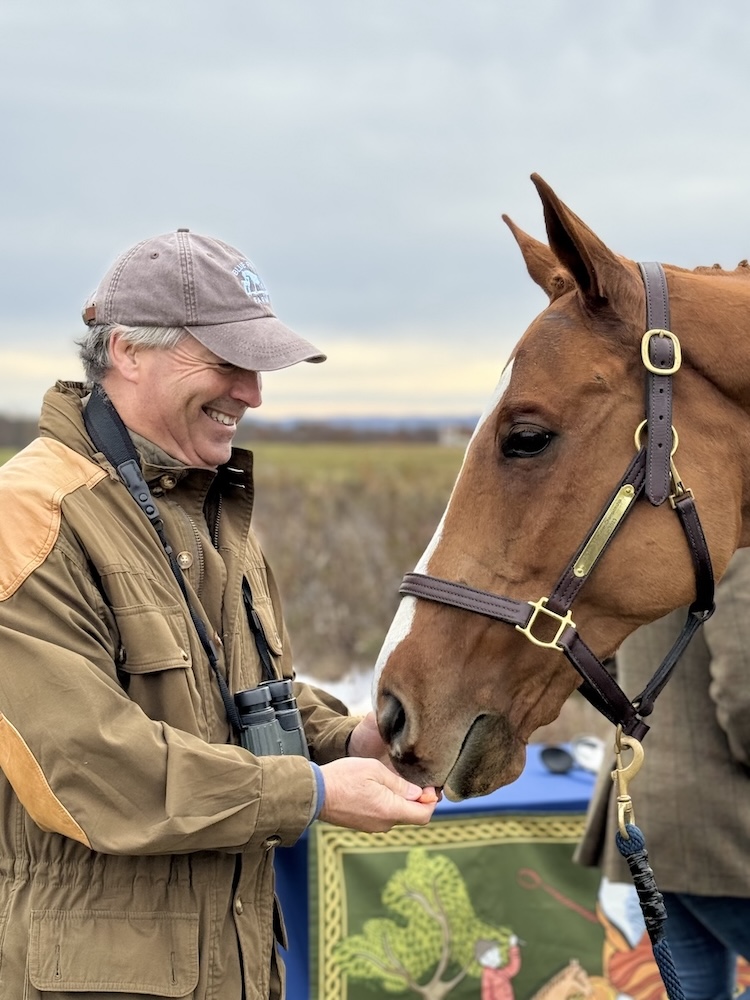 A smiling man feeding a chestnut horse.