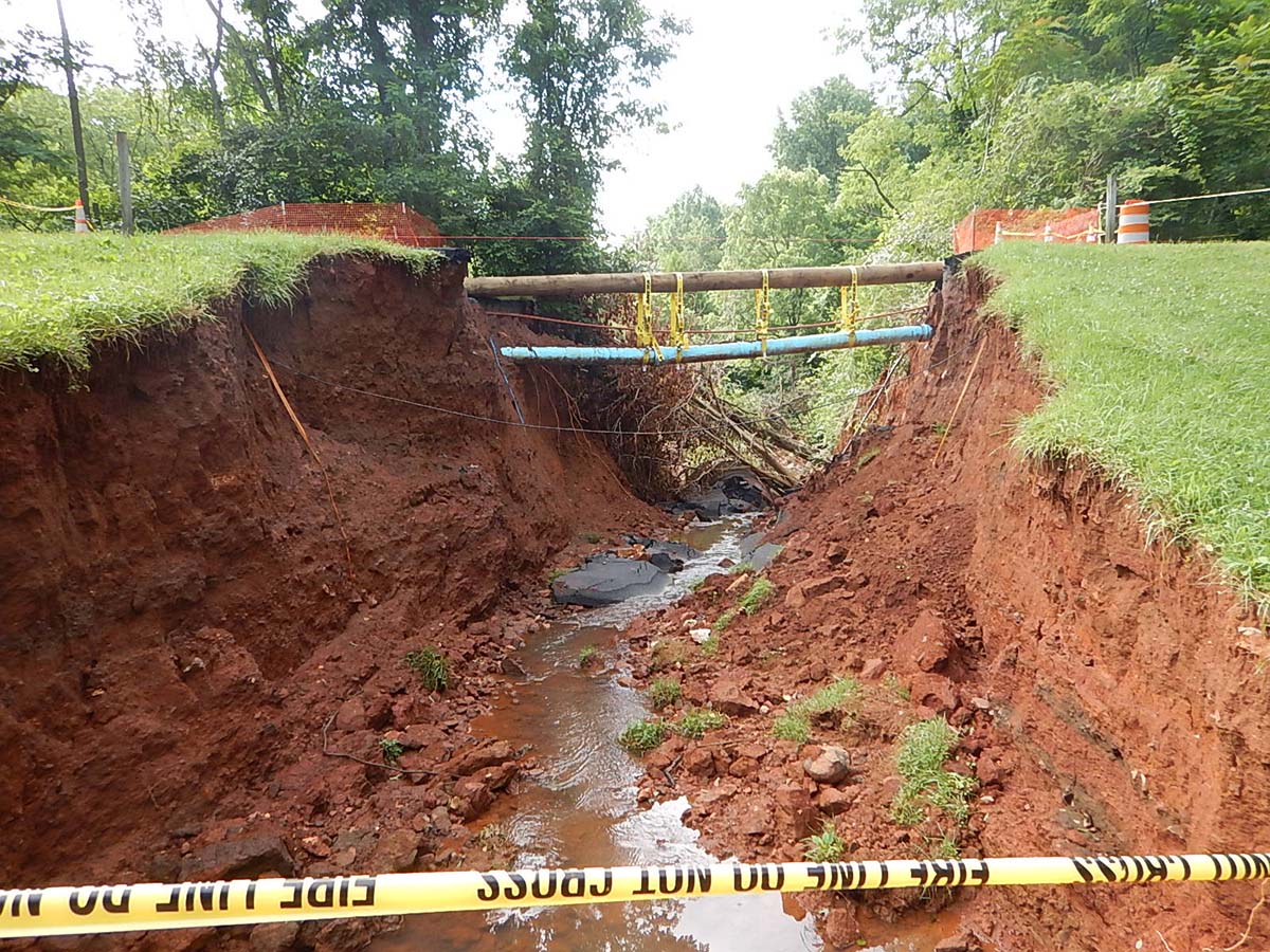 The land is washed away under a road, with pipes exposed and caution tape lining the sides.