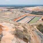 aerial view of an open-face dirt hills of an open pit gold mining, with green/brown tinted retention ponds and construction vehicles.