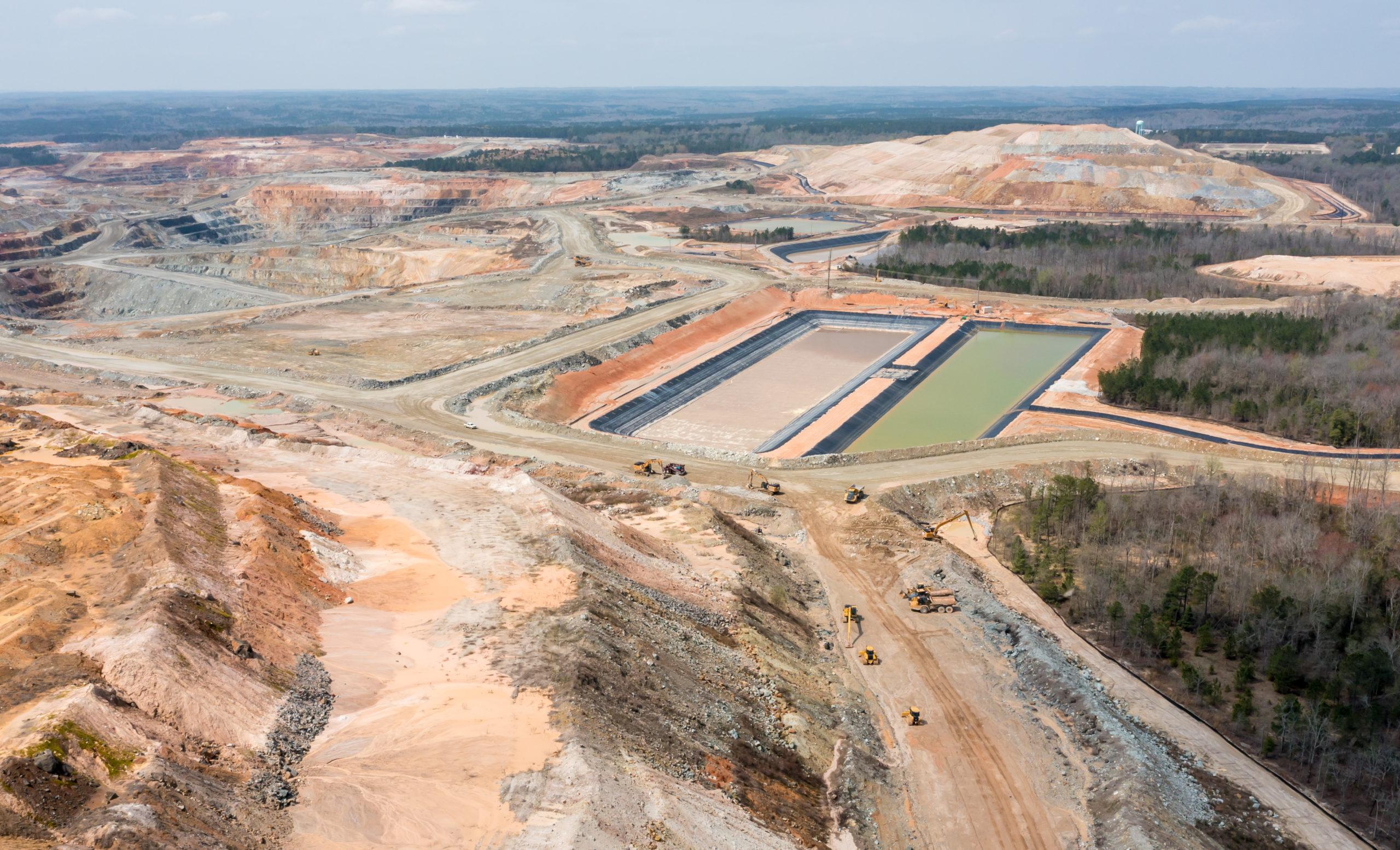 An aerial view of an open-face dirt hills of an open pit gold mining, with green/brown tinted retention ponds and construction vehicles.