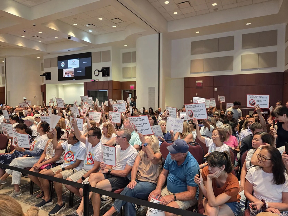 Crowd of protesters seated in a meeting hall holding up signs.