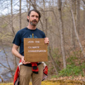 a man stands with trees behind, presenting a wooden sign that says "Join the climate conversation"