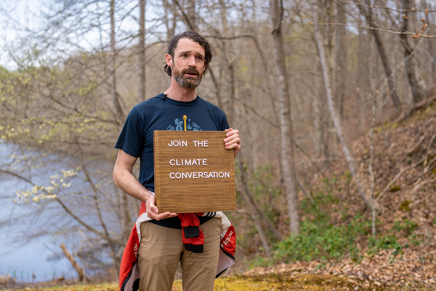 a man stands with trees behind, presenting a wooden sign that says "Join the climate conversation"