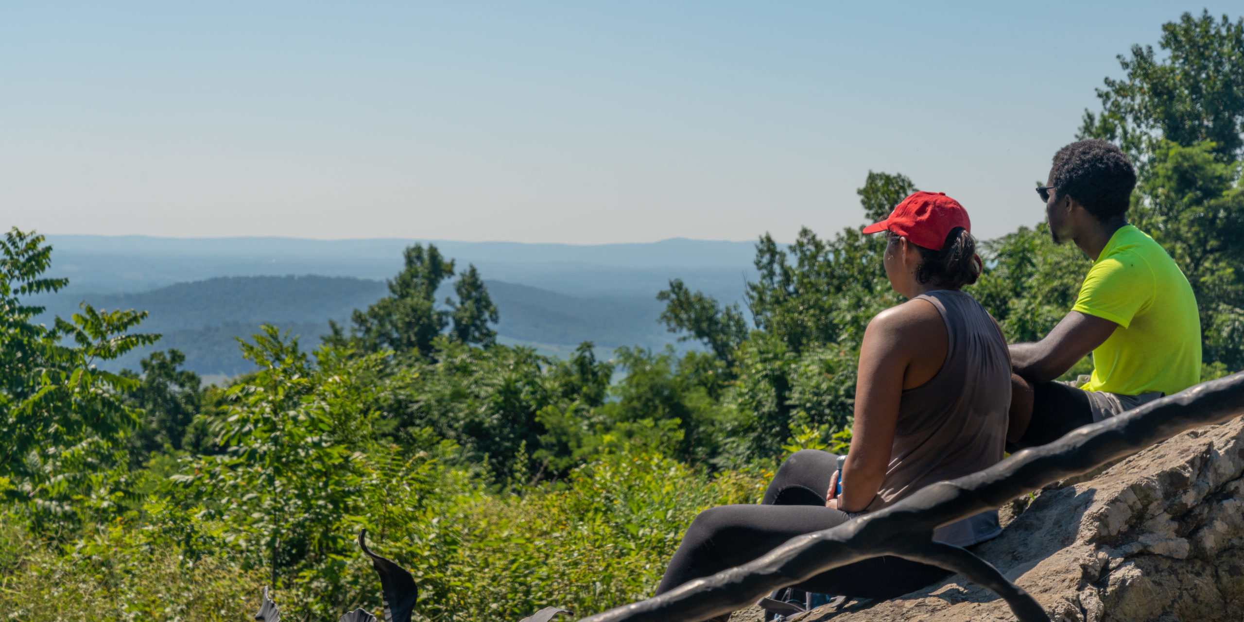 two young people sitting on rocks looking at a valley of blue mountains during the summertime