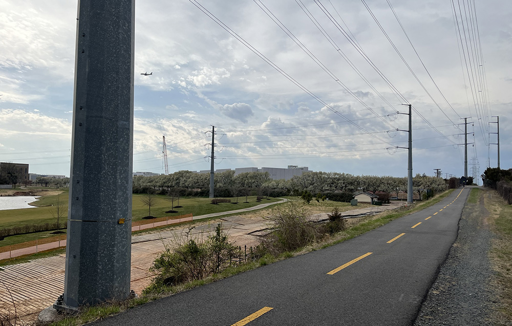 A biking path with large gray metal transmission line poles right next to it at regular intervals going into the distance and then to the left, where they pass in front of a data center under construction.