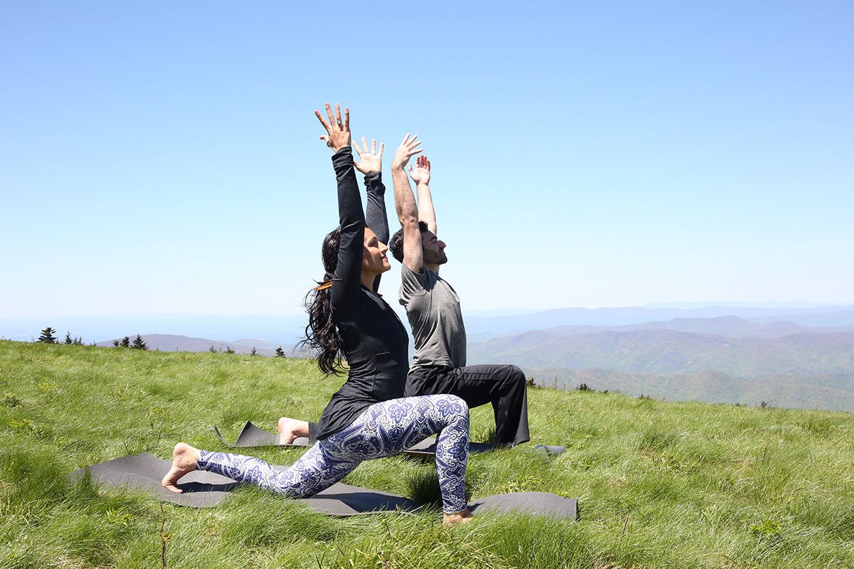 A man and woman wearing exercise attire practice yoga on yoga mats in the grass, with mountains in the background.