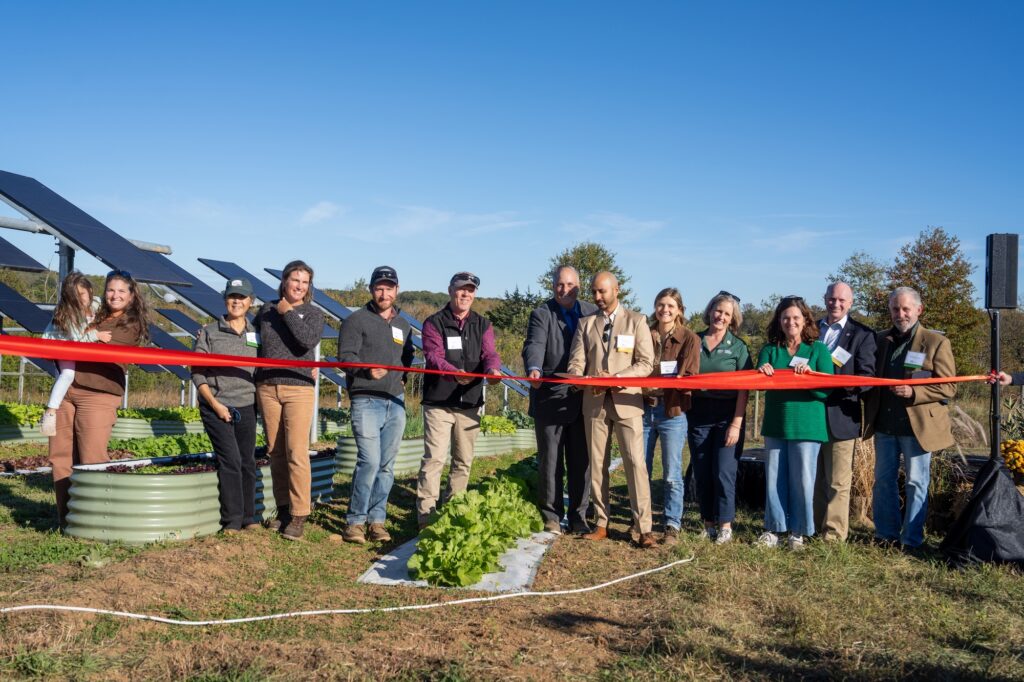 A row of smiling people stand behind a red ribbon in front of solar panels and crops, about to cut the ribbon with ceremonial scissors.