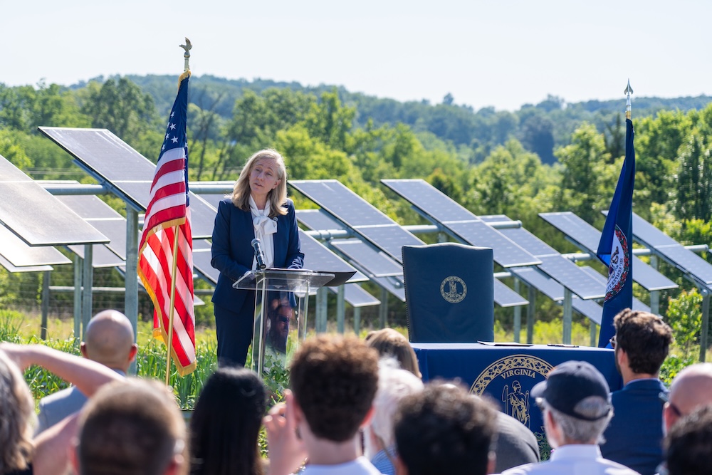 A woman at a podium, in front of a field of solar panels and framed by an American flag and a Virginia state flag, addresses a crowd.