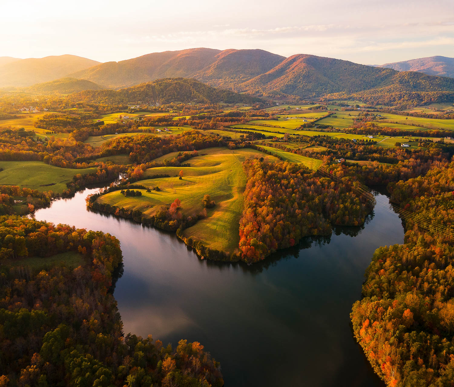 sunset falls over a reservoir surrounded by autumnal foliage and green pastures, with mountains on the horizon