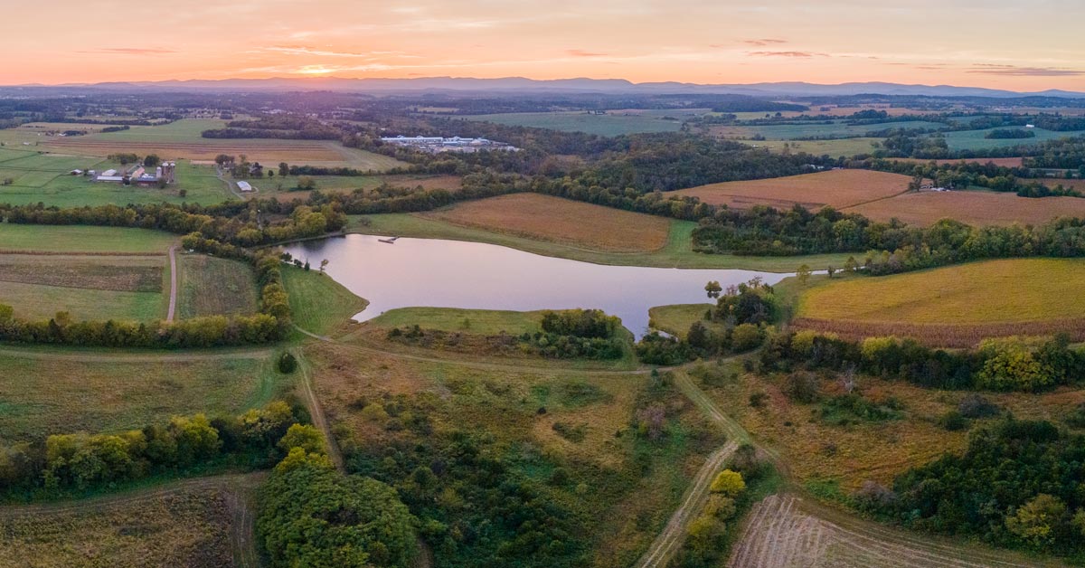 aerial photo of fields, trees and large pond with mountains and sunset in background
