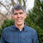 headshot of man with blue shirt with green shrub in background