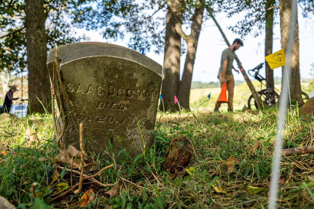 old gravestone on overgrown grass with person walking in the background