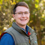 Young man wearing glasses and a green vest smiles against a leafy green backdrop.