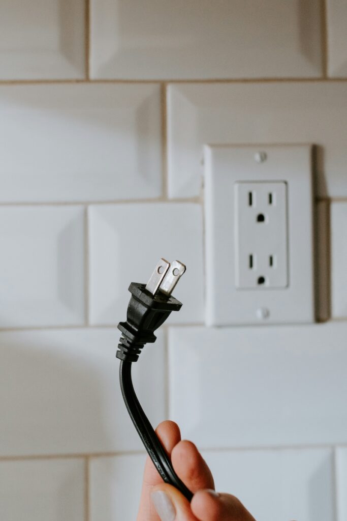 A hand holds an unplugged power cord in front of an electrical outlet.