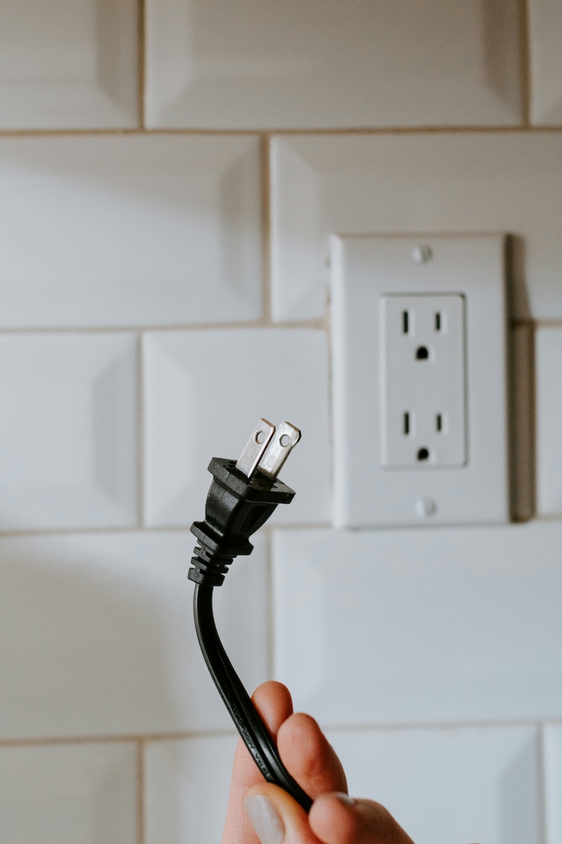A hand holds an unplugged power cord in front of an electrical outlet.