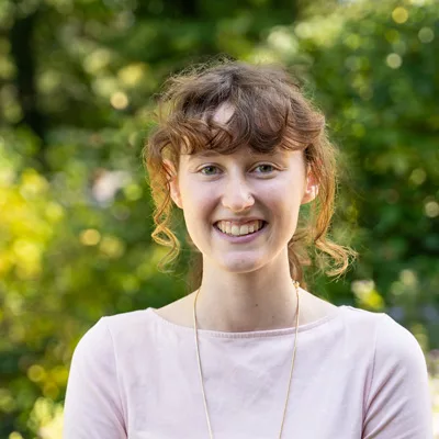 headshot of woman with brown hair and a pink shirt with greenery behind her