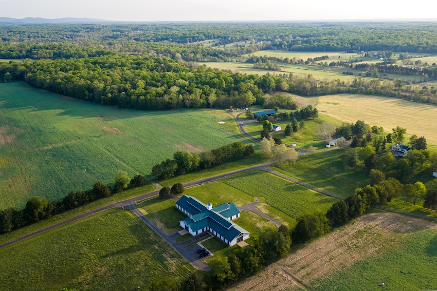 An aerial view of a large horse barn surrounded by green fields and forest.