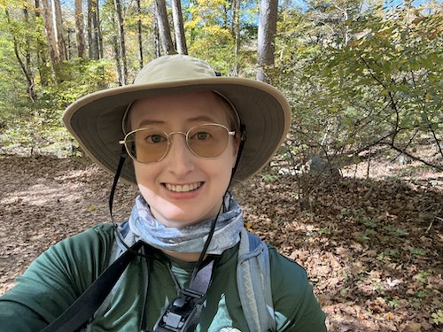 A selfie of someone in the woods, wearing a backpack and sunhat.