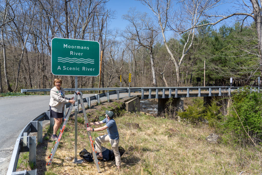 Two PEC employees stand on a ladder under the Moormans River - A Scenic River sign.