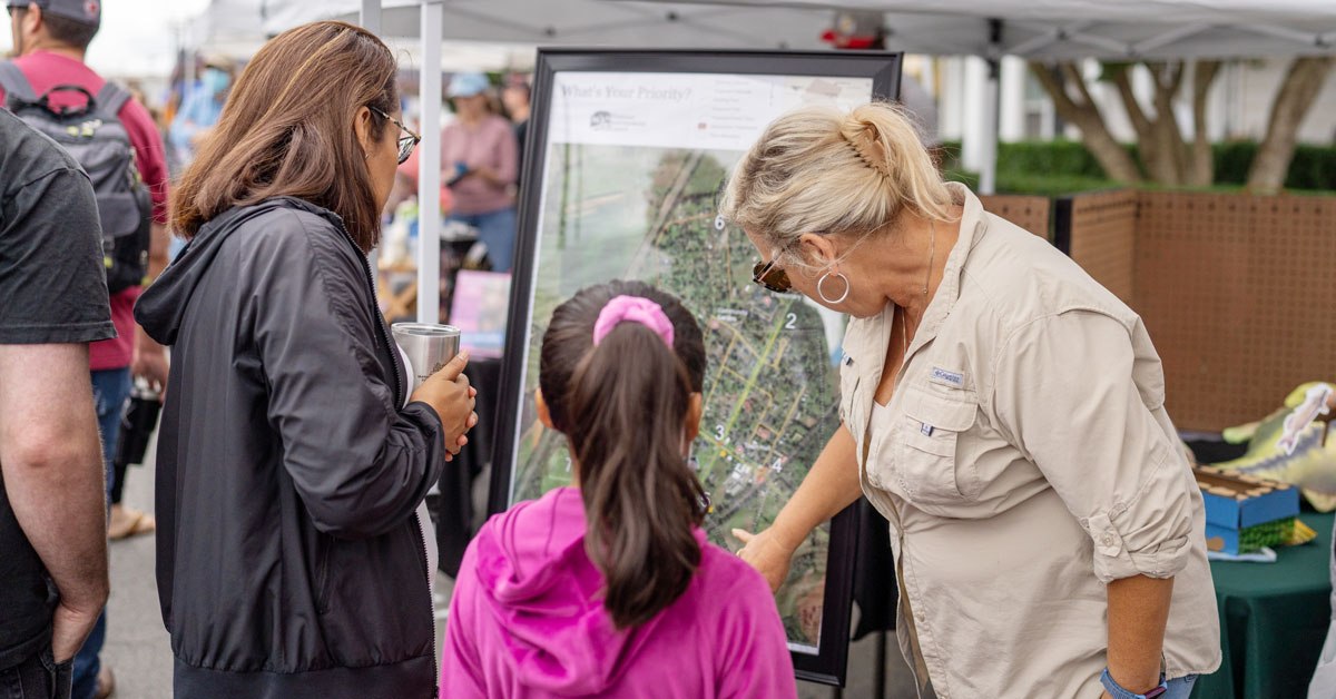 woman in tan shirt talking to a woman in a black jacket and child in a pink sweatshirt. She's pointing to a map on stand near a table under a canopy.