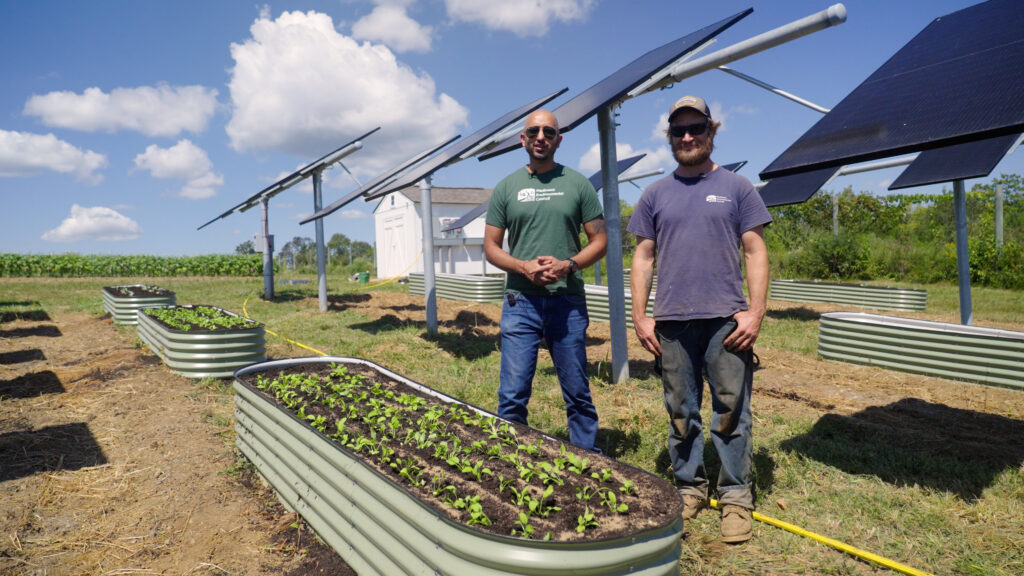 Ashish Kapoor and Teddy Pitsiokos stand in front of a raised bed with ground-mounted solar panels behind them.