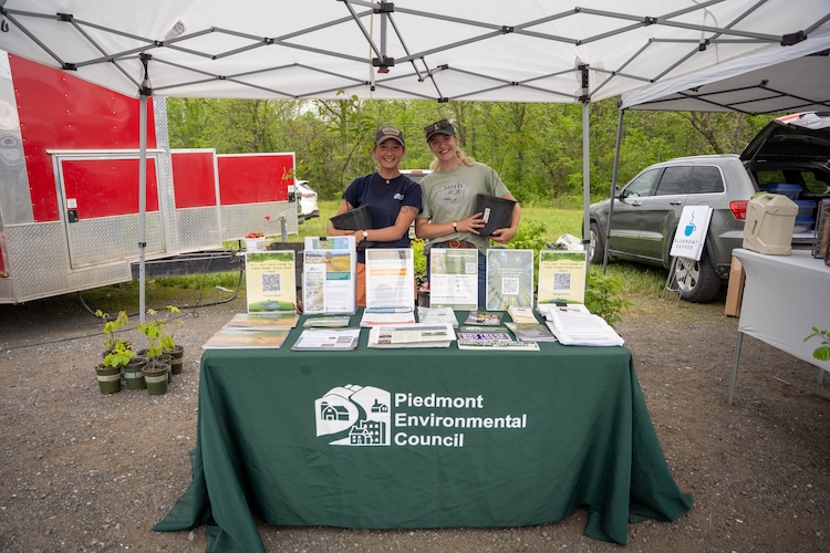 Two people stand behind a table holding pots with tree saplings. The table is covered in informational flyers and the front reads "Piedmont Environmental Council."