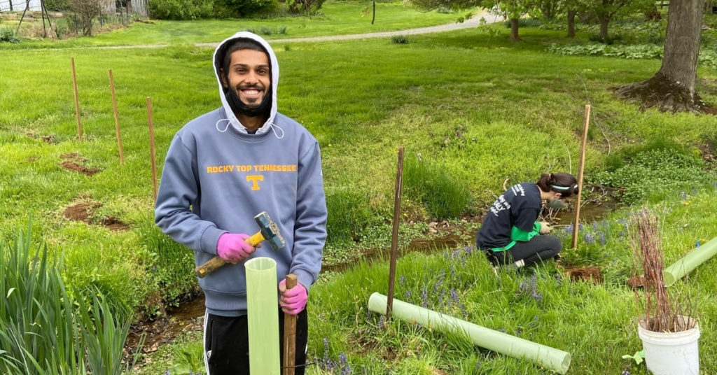 Young smiling man hammering a tree tube into the ground next to a stream