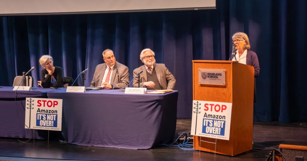 woman speaking on stage at podium beside a blue table with three other presenters who are sitting down