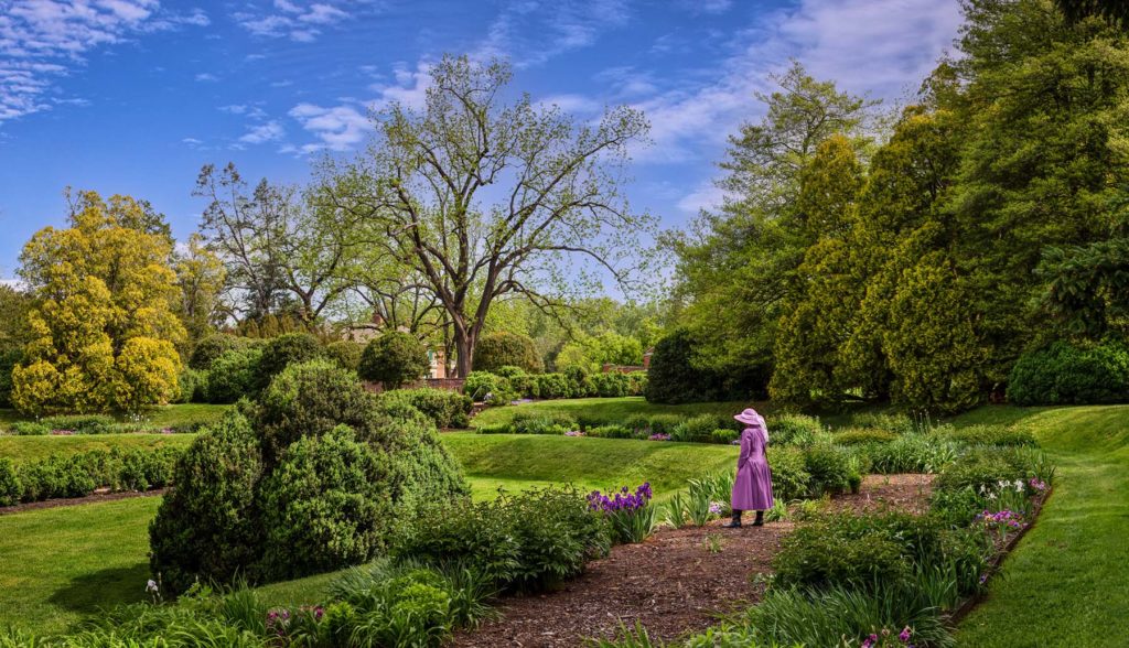 A woman walks in a garden during spring, dressed in a pink dress and bonnet